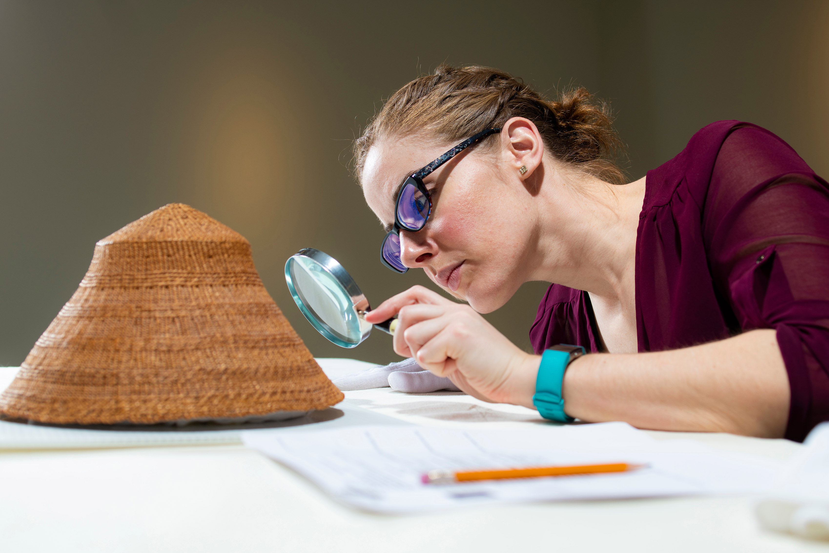 UAlberta Museums staff closely examines a wicker hat with a magnifying glass