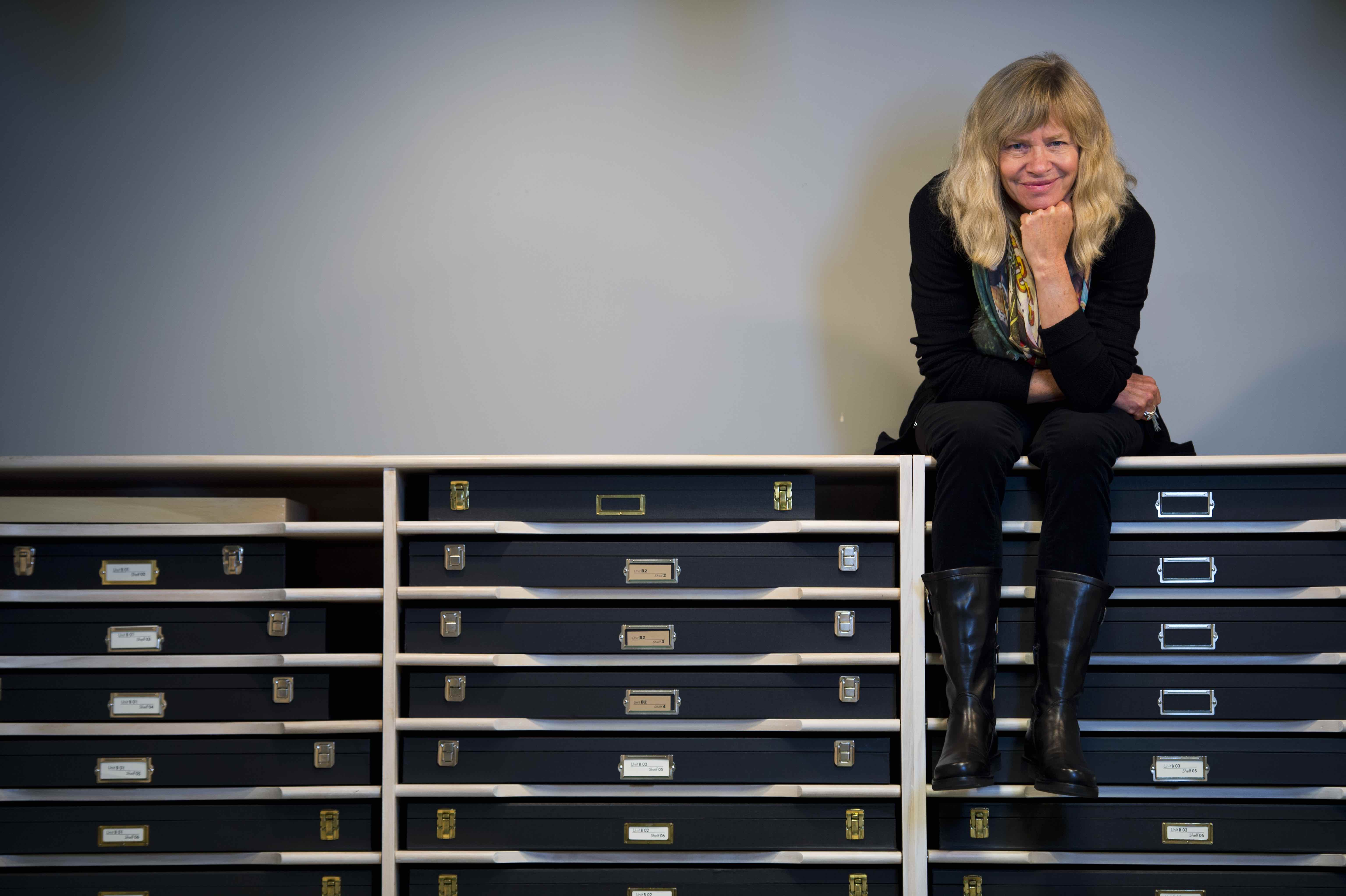 Woman sits on top of a shelving unit holding boxes. 