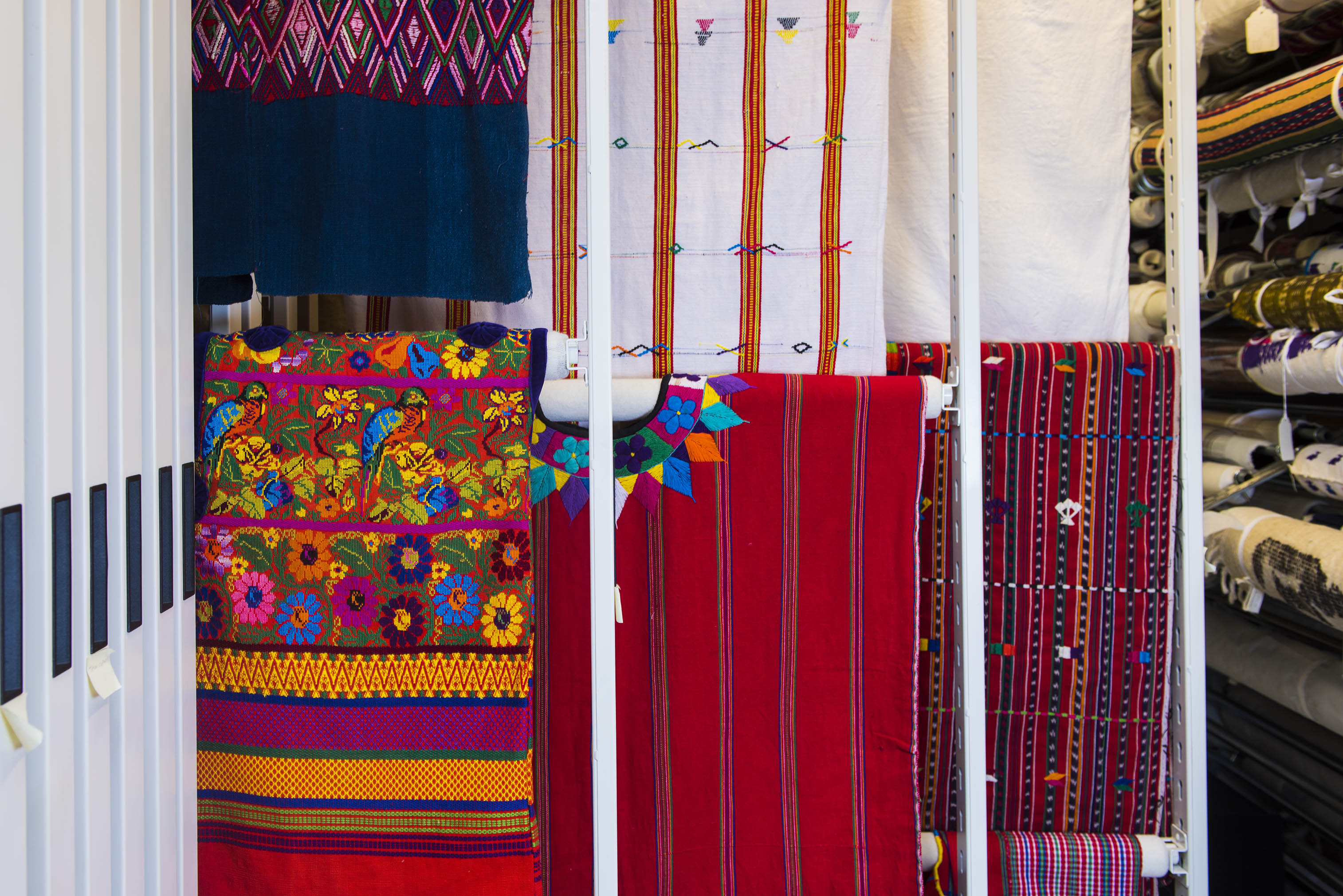 Two rows of colourful textiles vertically stored on padded, horizontal rods that extend from a shelving unit.