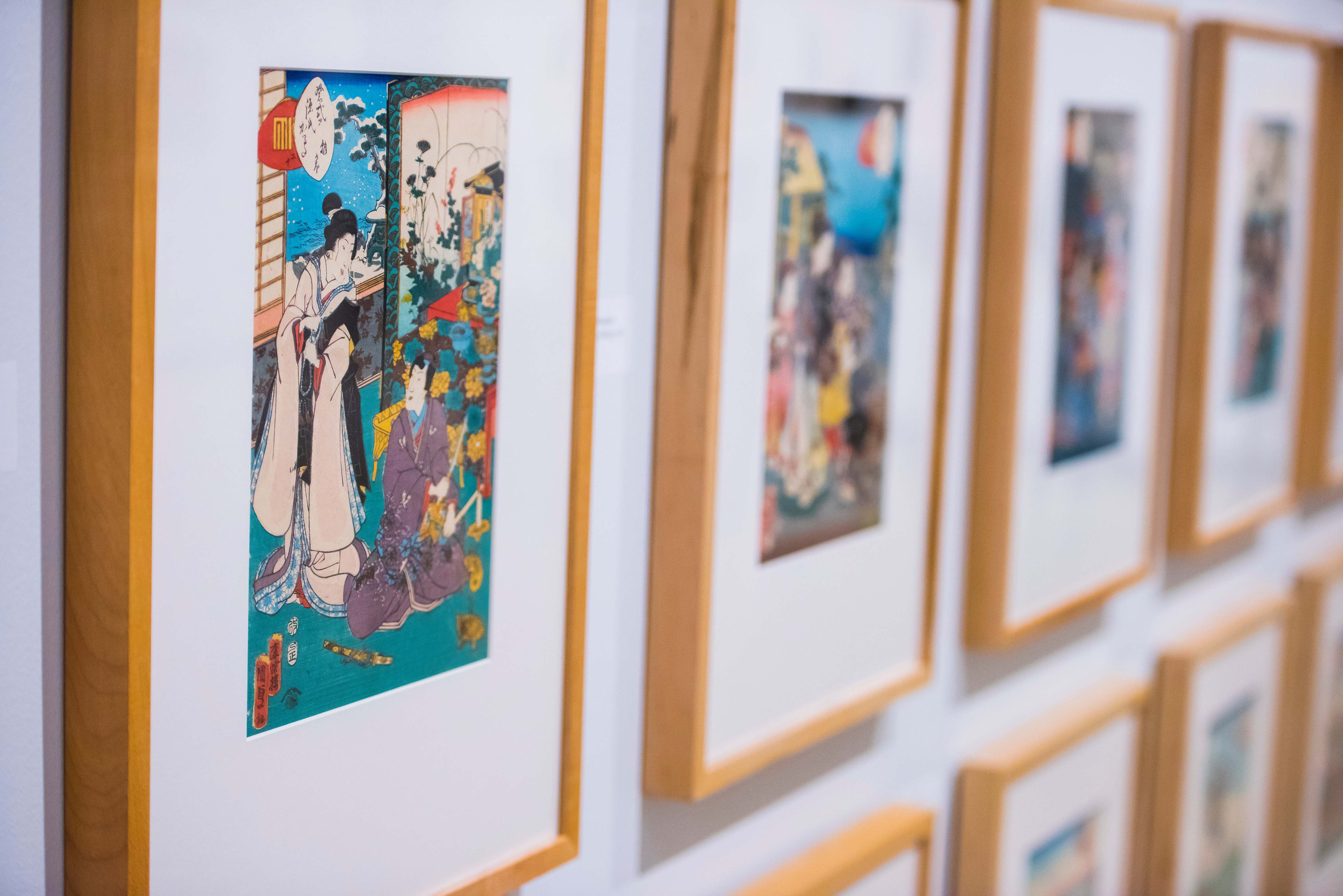 Angled perspective of colourful Japanese illustration prints that are hung in small light wooden frames on a white wall.