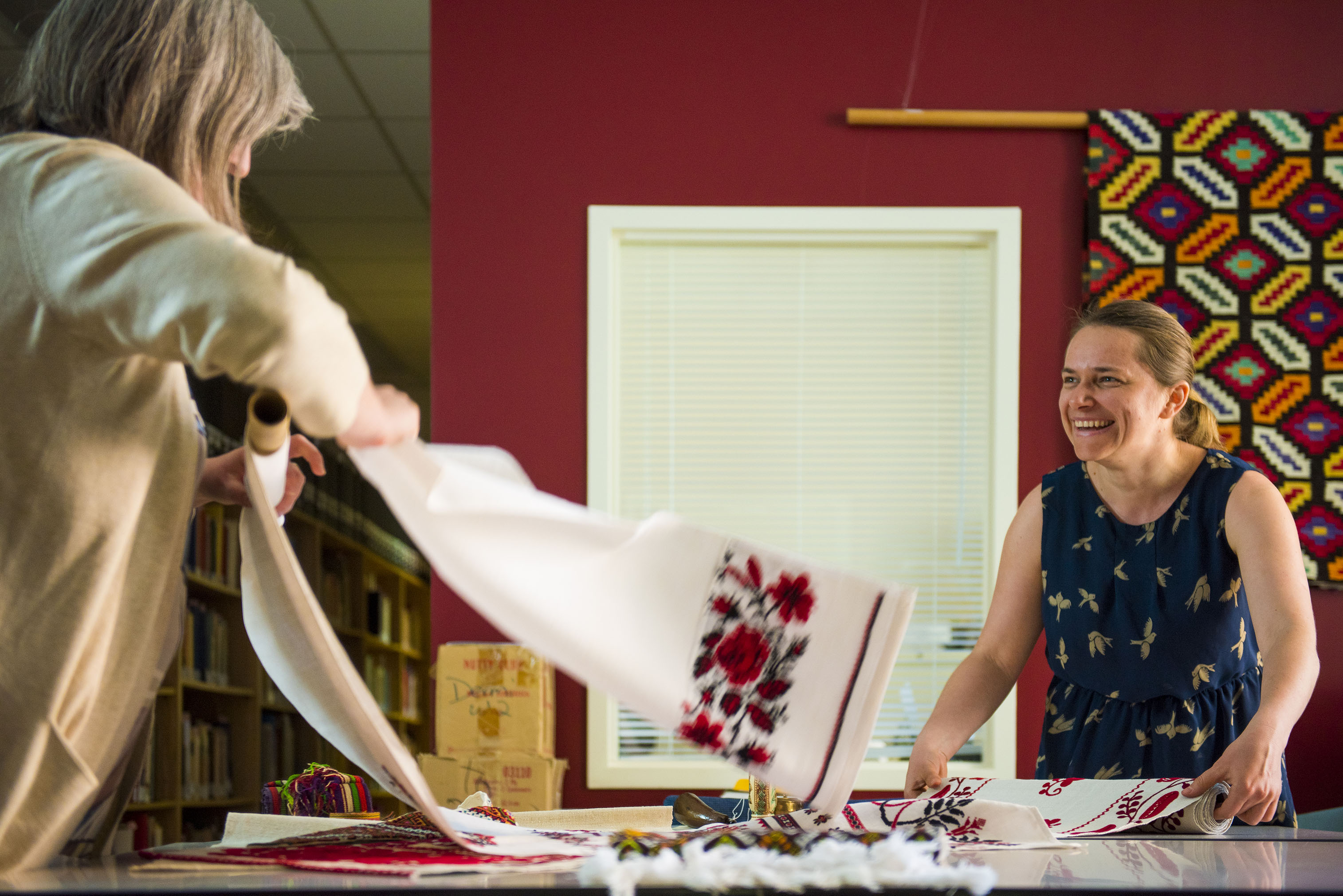 Two volunteers laugh while rolling white textiles with intricate red embroidery designs