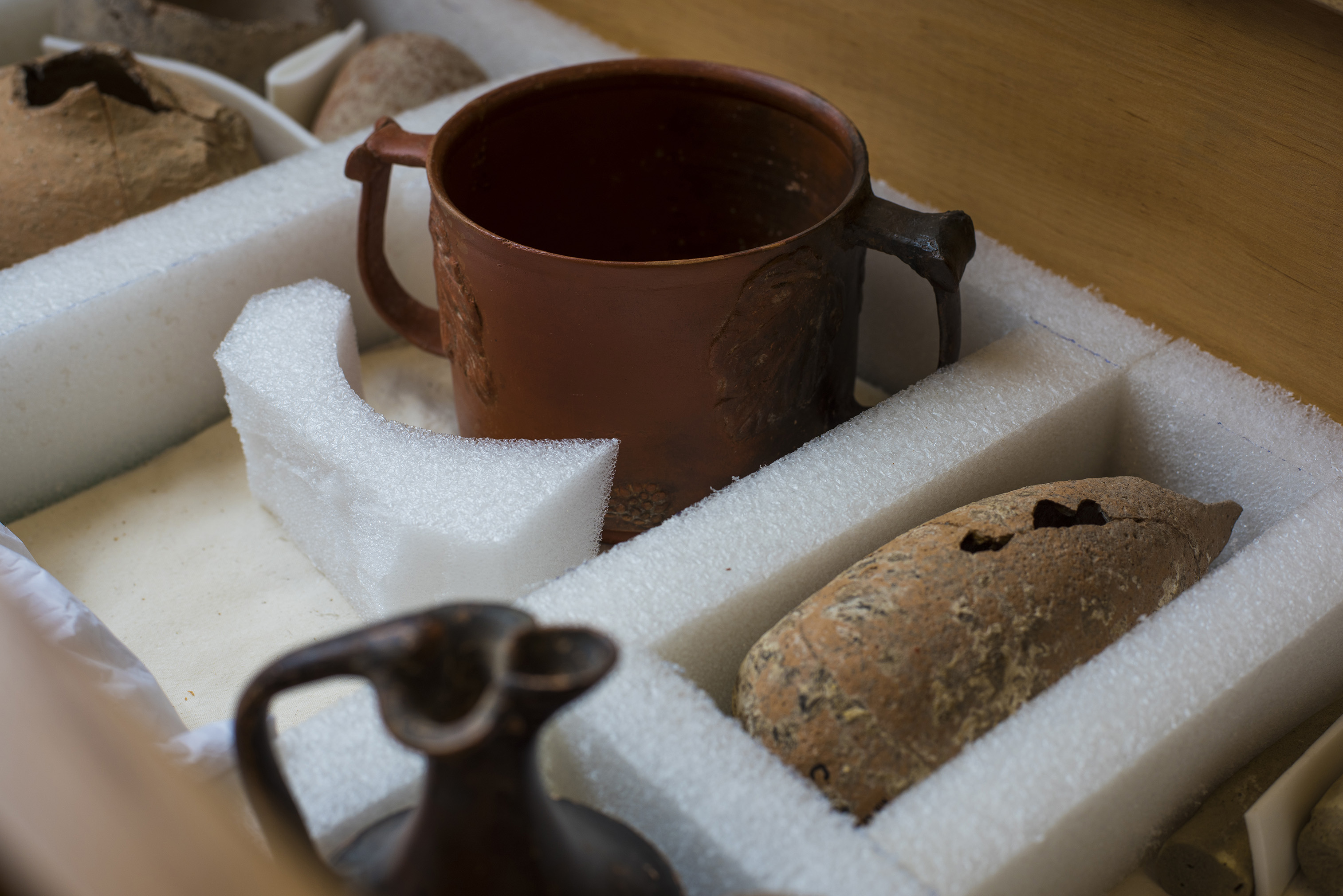 A red-brown, two-handled vessel sits nestled between foam pieces, surrounded by other small fragmented vessels.