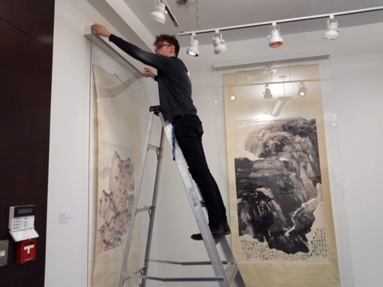 Martin stands on a ladder, adjusting a plexiglass frame covering a scroll.