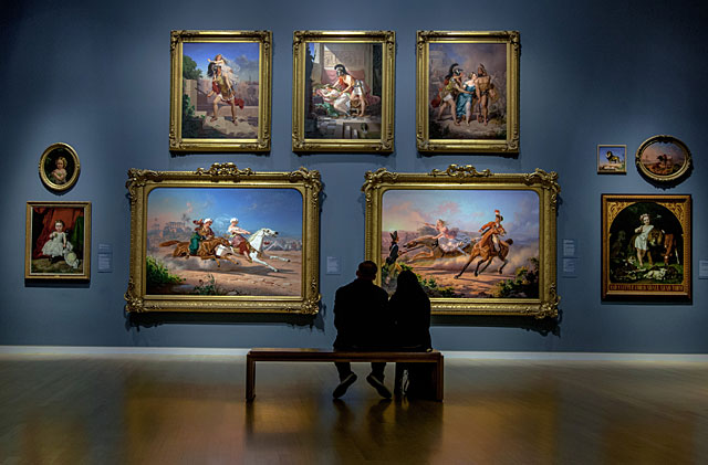 Two individuals sitting on a bench in a gallery, looking at a wall of oil paintings
