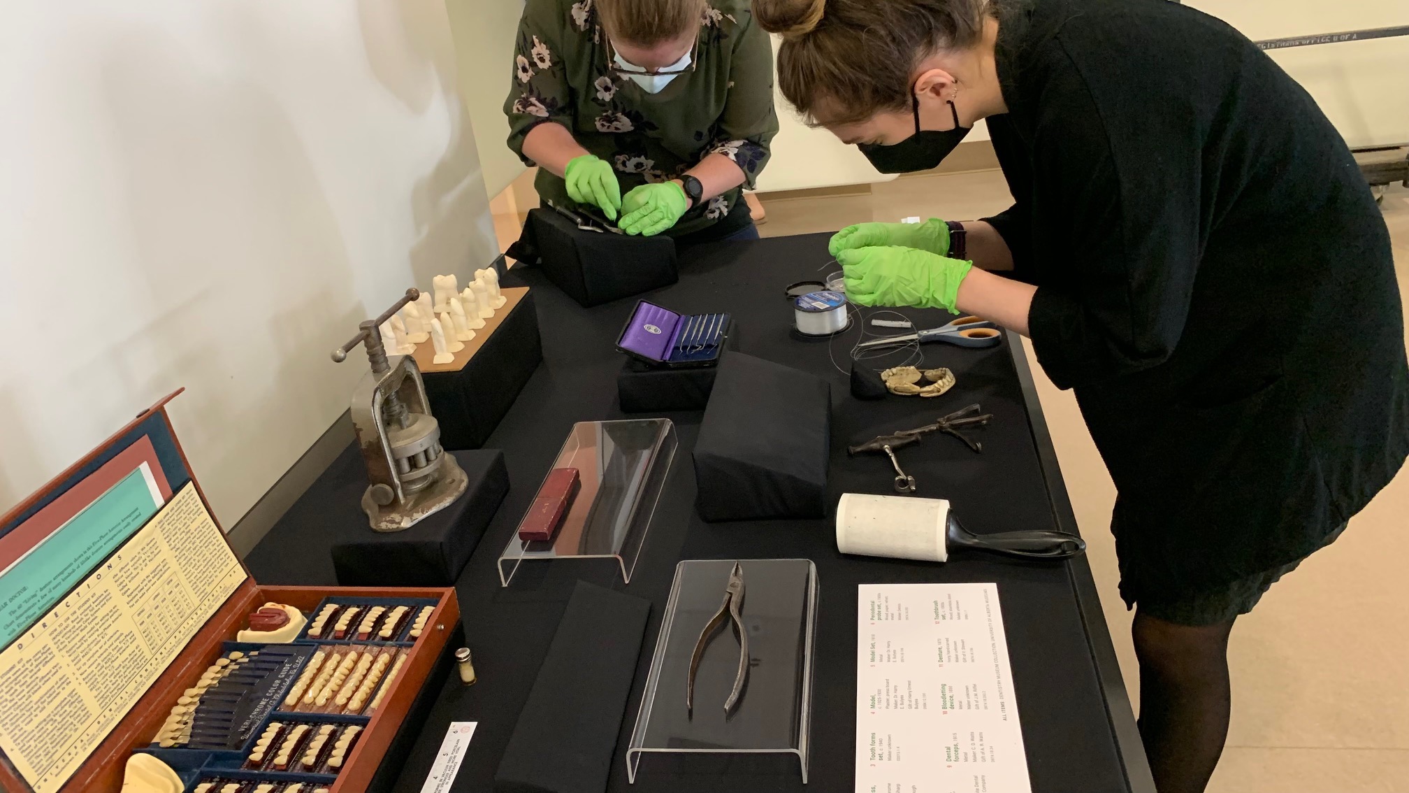 Two white women wearing green nitrile gloves arrange dental instruments for a display case on a black fabric-covered table. 