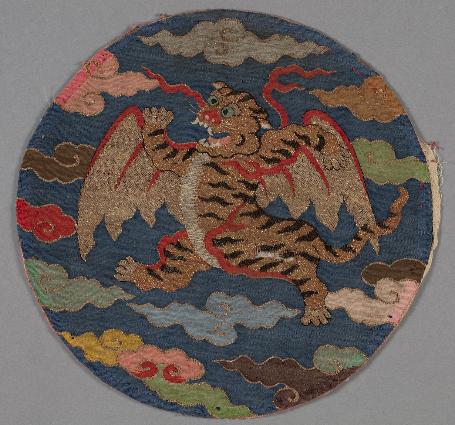 A round textile has a tiger centred in the design with wings of gold outstretched. The tiger, with teeth exposed, is in an active pose and is surrounded by a blue background and different coloured swirling clouds.