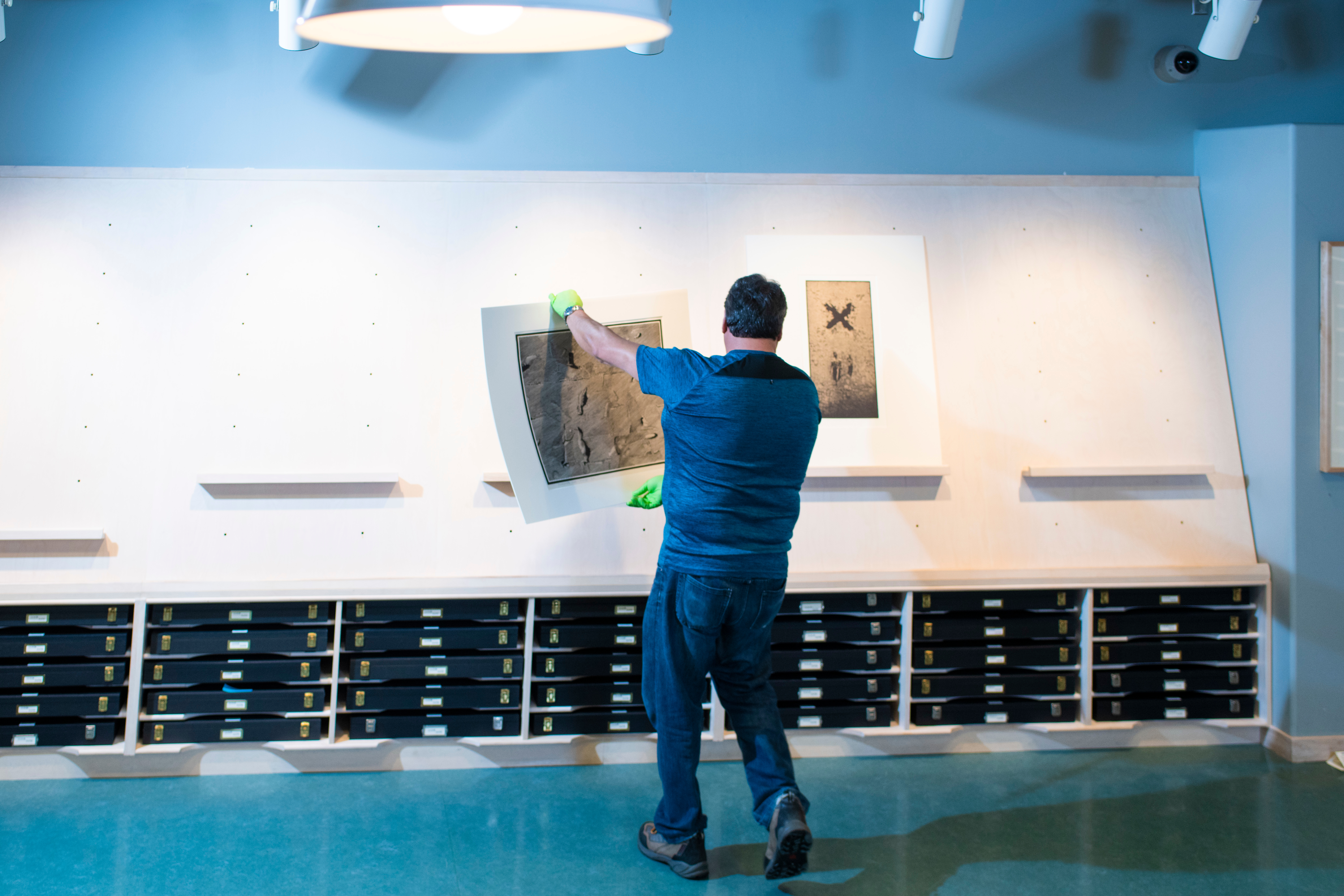 A man wearing a blue shirt and jeans stands facing towards a wall with rows of black boxes. In his arms, he holds a work of art and moves to place it leaning against the wall.