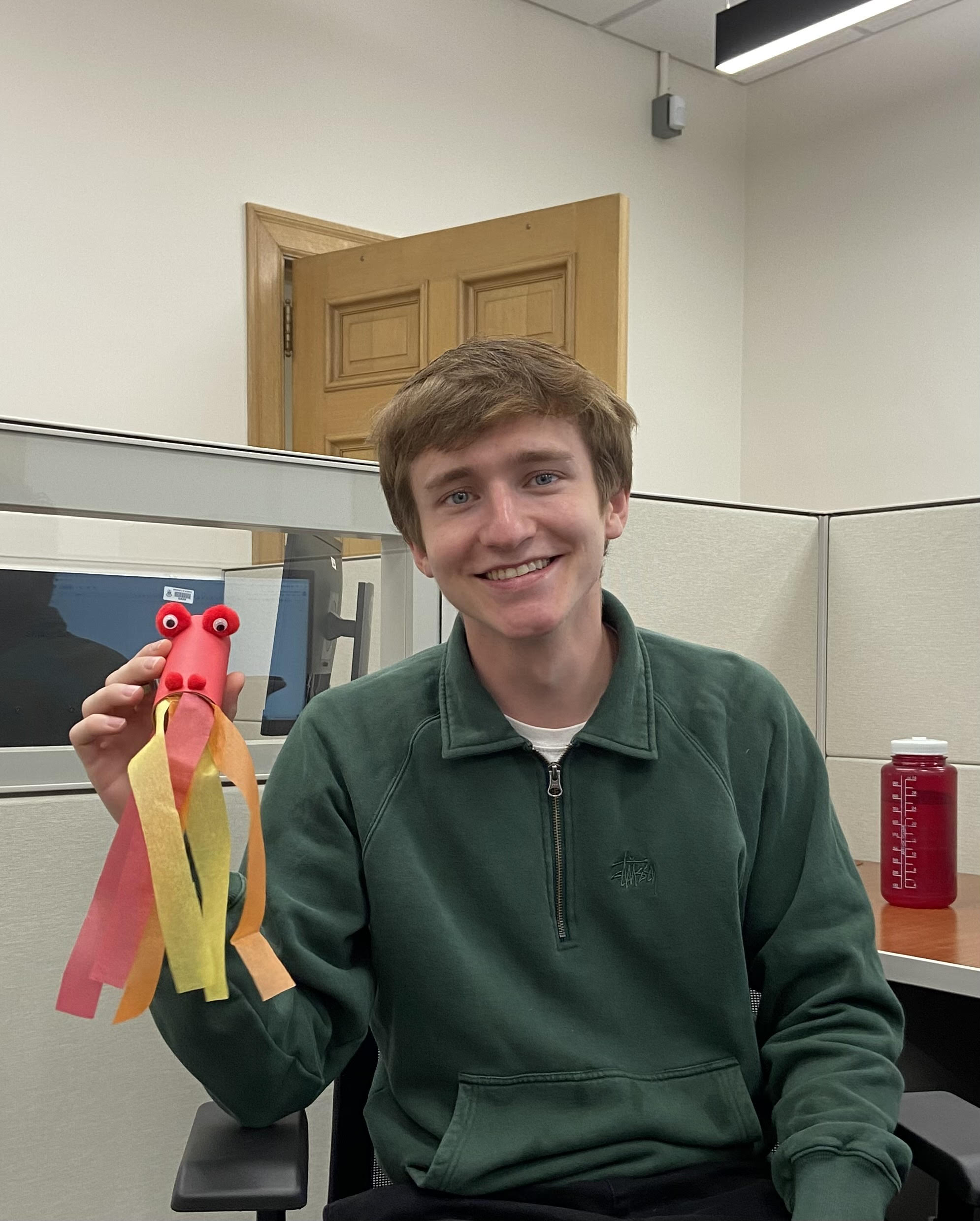 A smiling man in a green shirt, seated in a cubicle, holds up a dragon’s head crafted from colourful tissue paper.