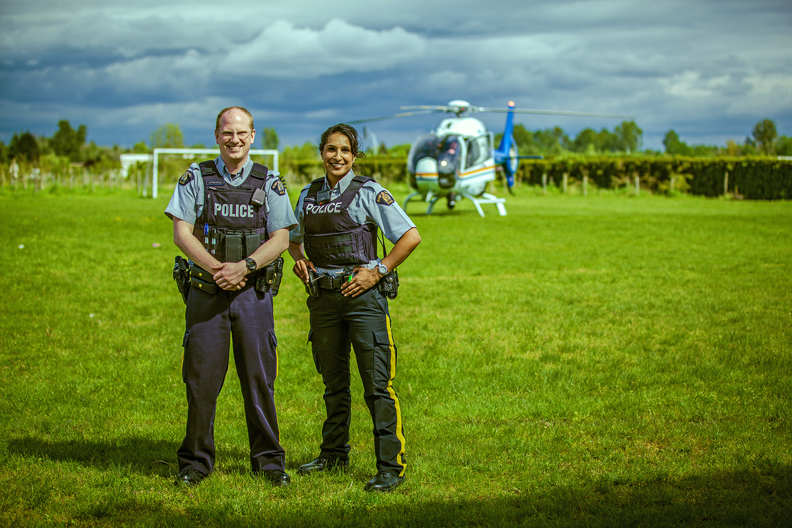 Image of two Edmonton Police Officers in the foreground smiling. There is a helicopter landed and out of focus in the background