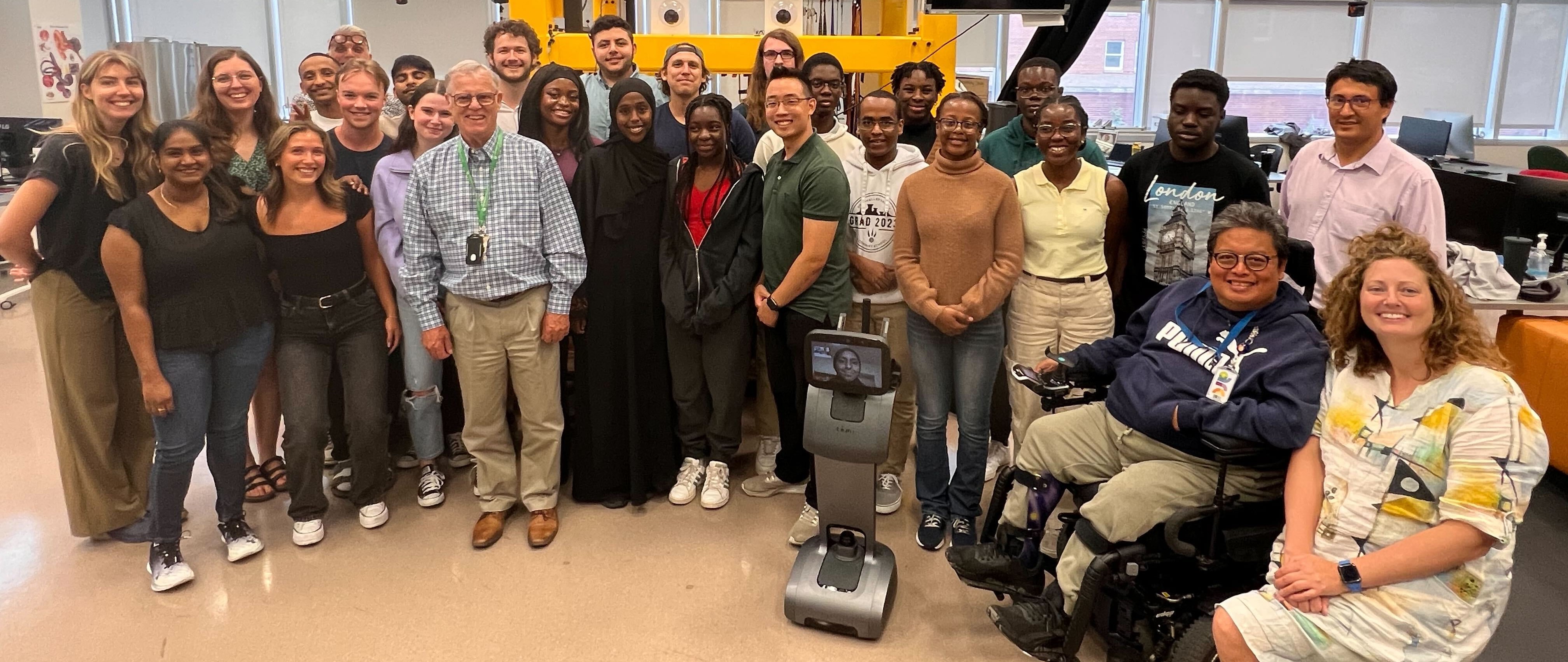 A wide photo showing the Rehab Robotics Lab's team from Summer 2023. There are 28 people and one person on a telepresence robot, smiling and in general chaotic disarray. You can tell a professional did not pose them.