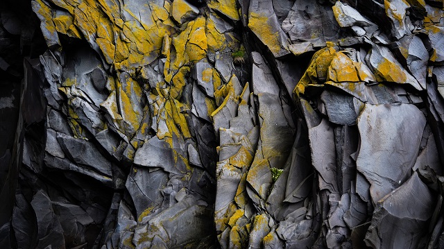 Closeup of a grey, jagged rock face with flecks of yellow discoloration and moss