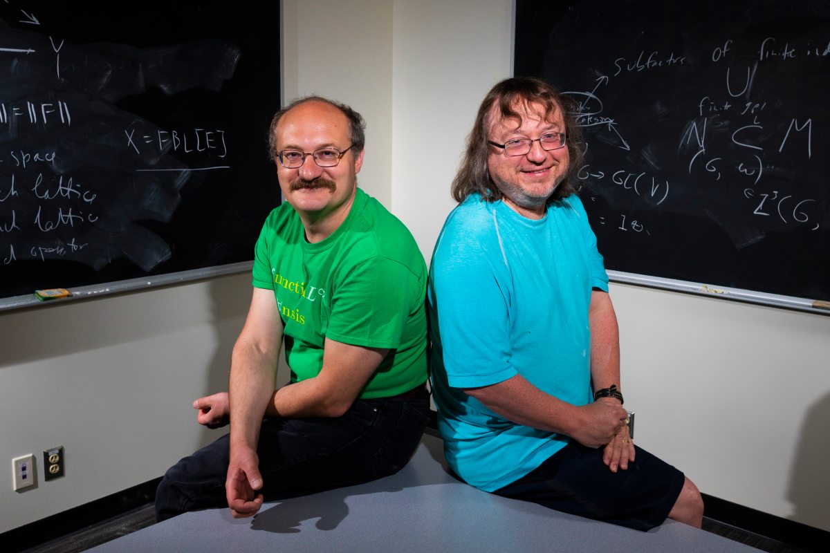 Math professors Vladimir Troitsky and Terry Gannon sit in front of two blackboards covered in equations.