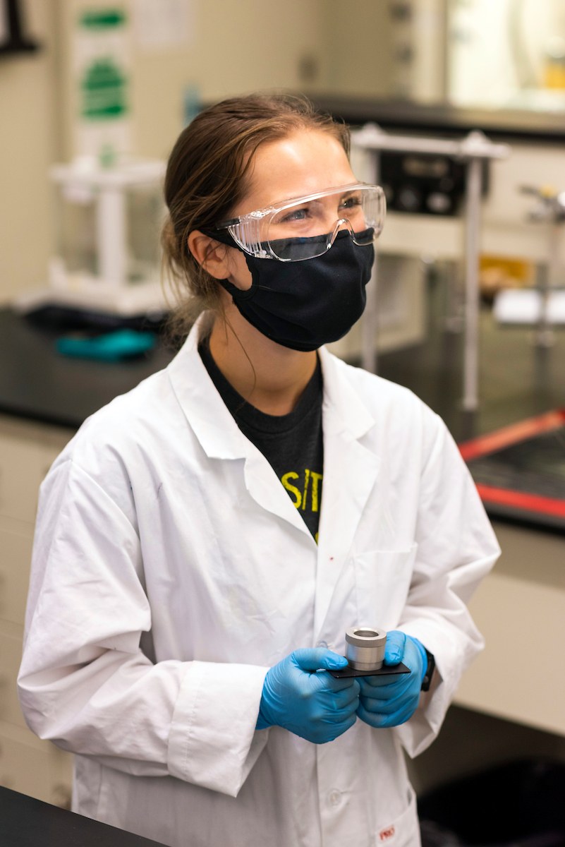 A smiling student works in an undergraduate chemistry lab while wearing a face mask and lab protective gear, including lab coat and goggles.