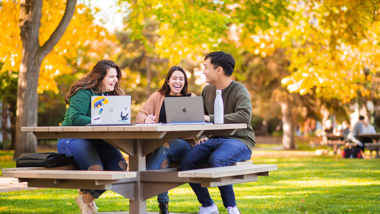 Three students sitting at a picnic table outside. The trees in the background have fall leaves coloured Green and Gold.