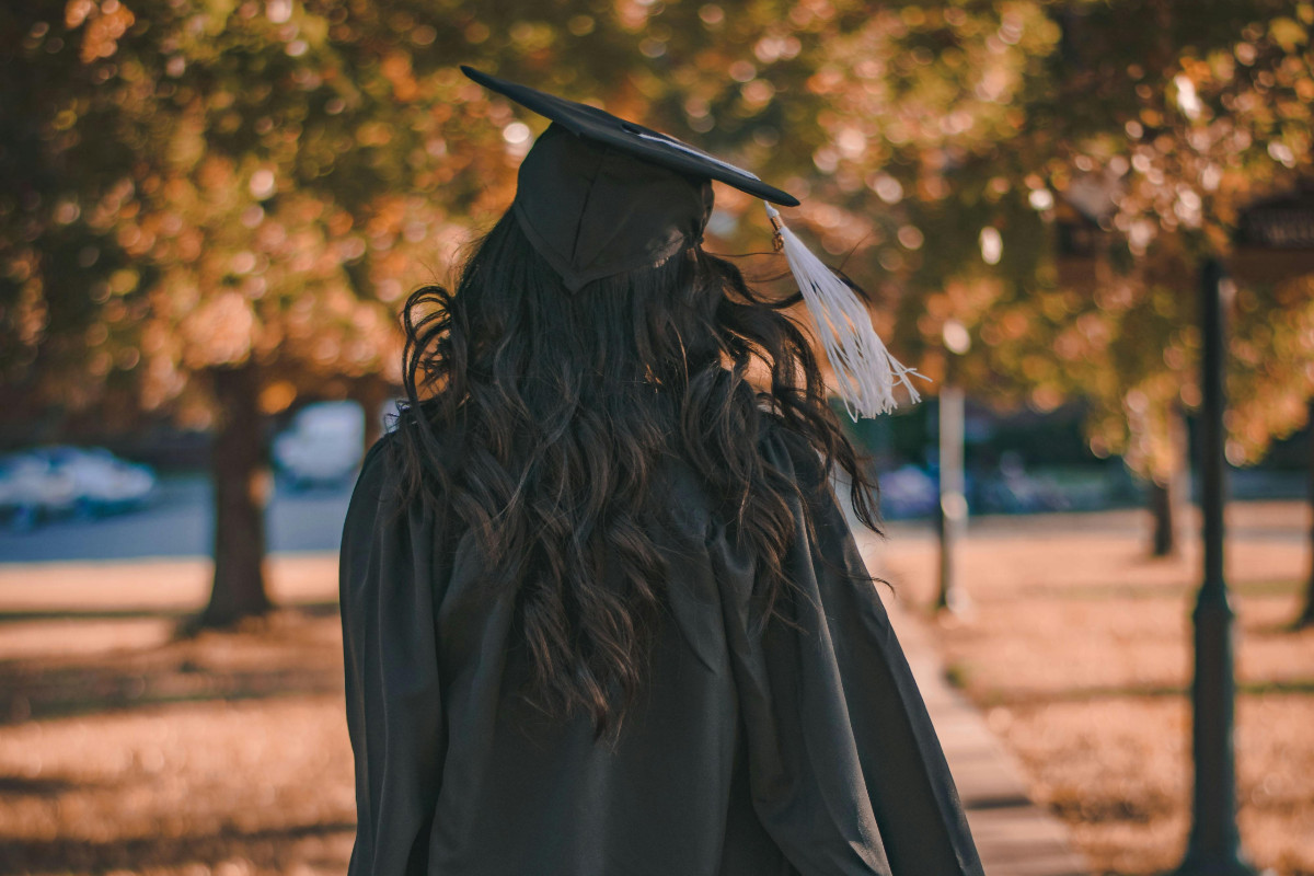 A student faces away from the camera. She is dressed in a black graduation cap and gown, pictured in a tree-lined quad.