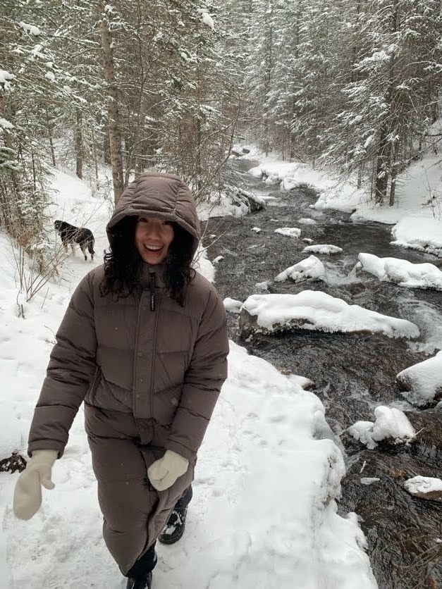 A smiling woman with long black hair in a brown parka and her dog walking by a stream in a snow-covered coniferous forest